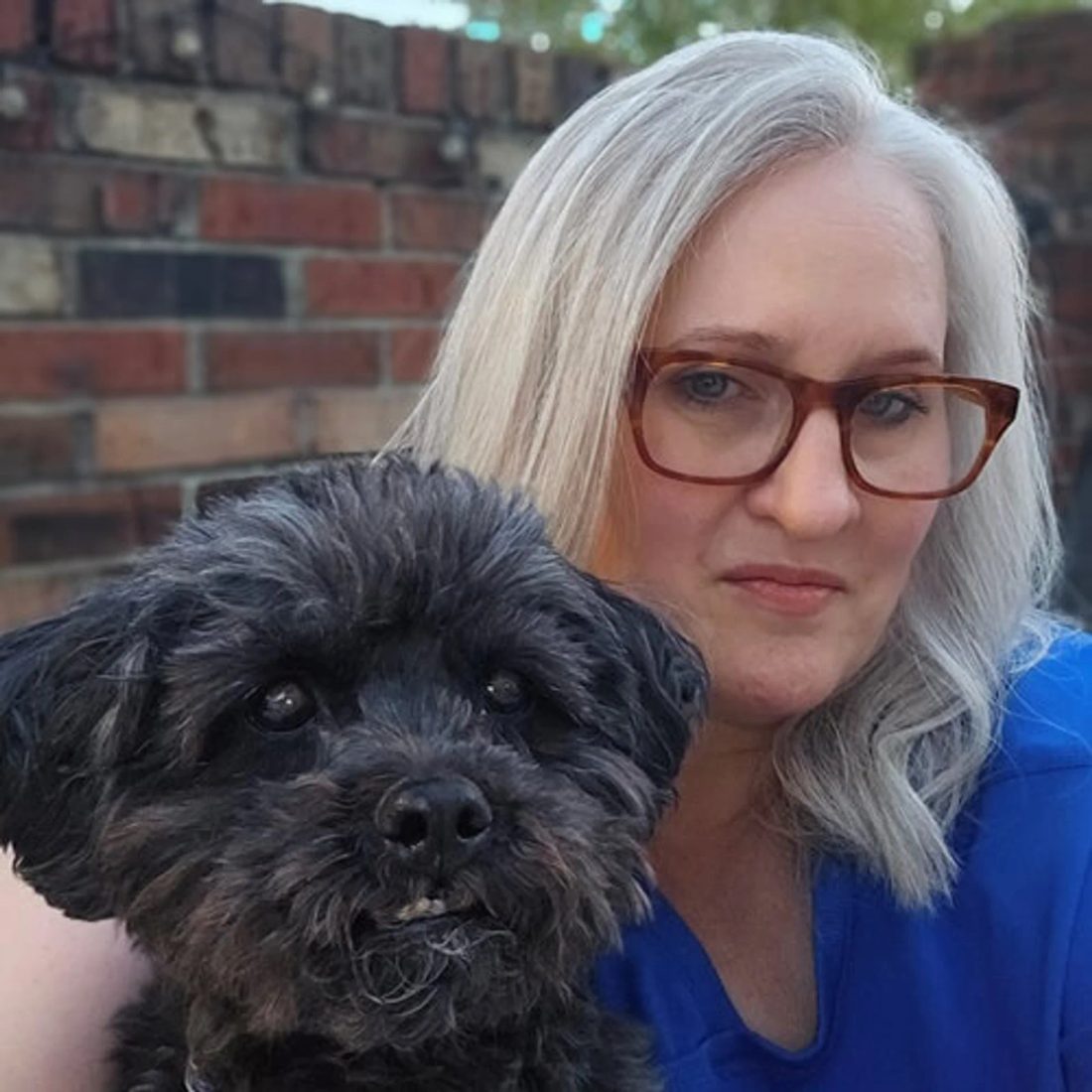 Woman with glasses holding a small black dog in front of a brick wall